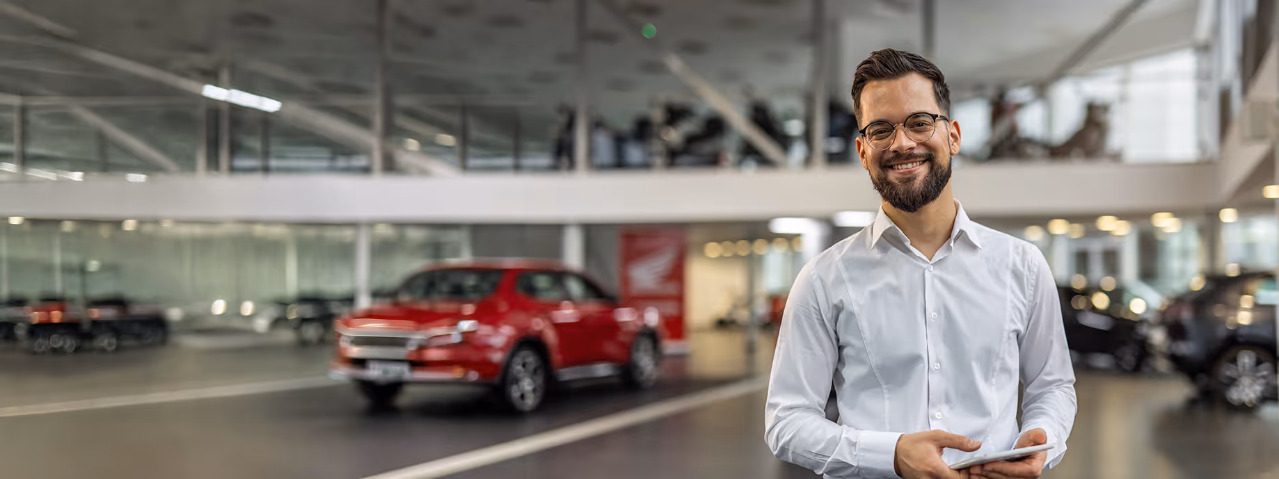 Mann mit Brille und weißem Hemd lächelt mit Tablet in einem Autohaus mit einem roten Auto im Hintergrund.