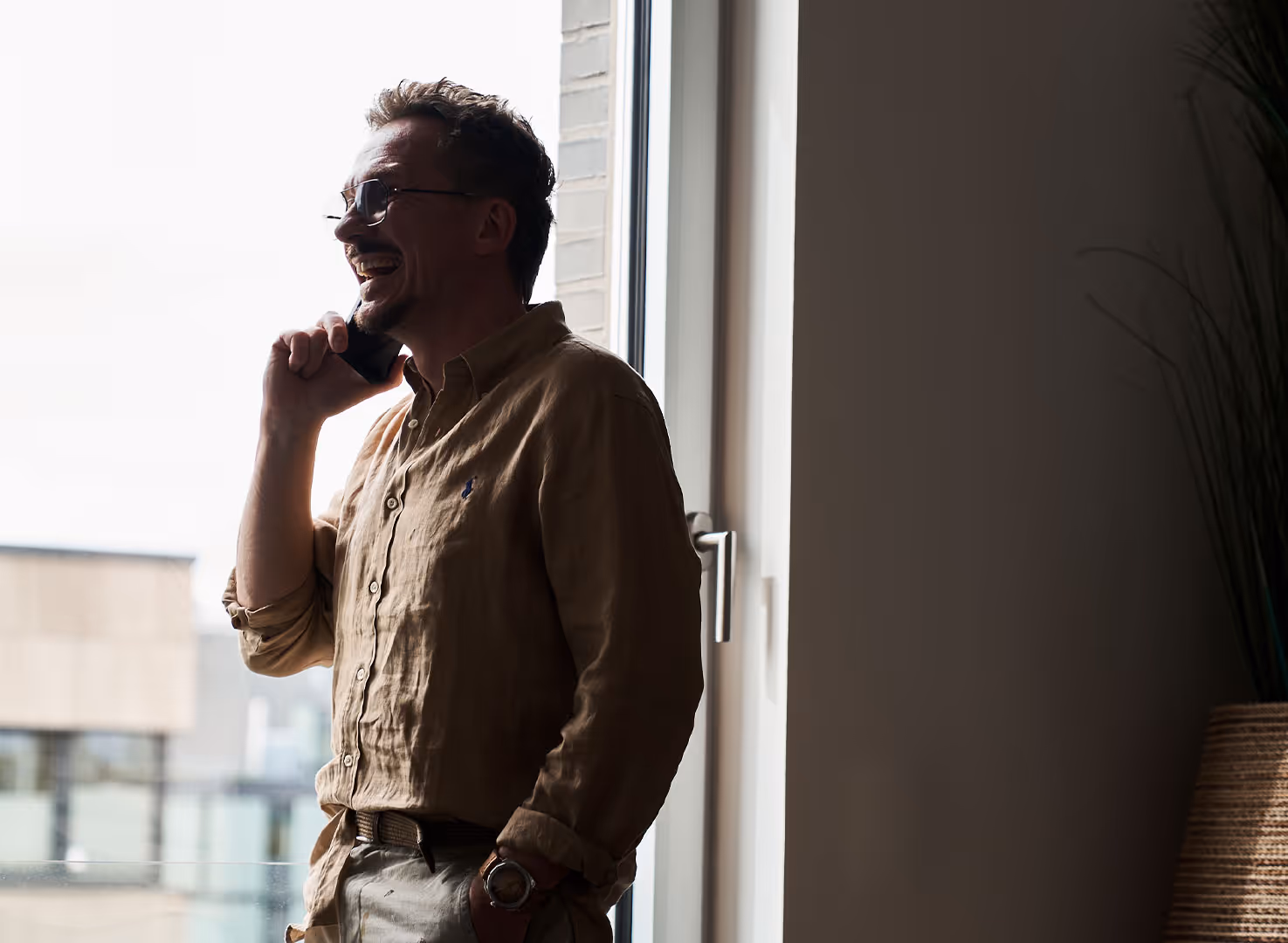 Lächelnder Mann mit Brille am Fenster telefoniert mit Handy, Hand in Hosentasche.