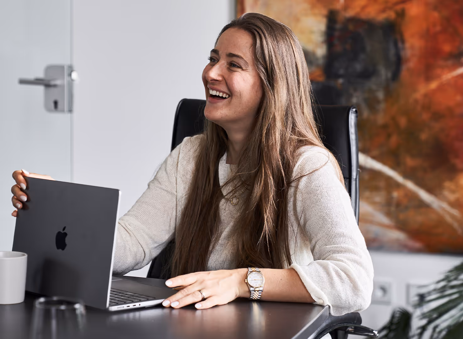 Frau sitzt lachend am Schreibtisch und hält einen aufgeklappten Apple Laptop.