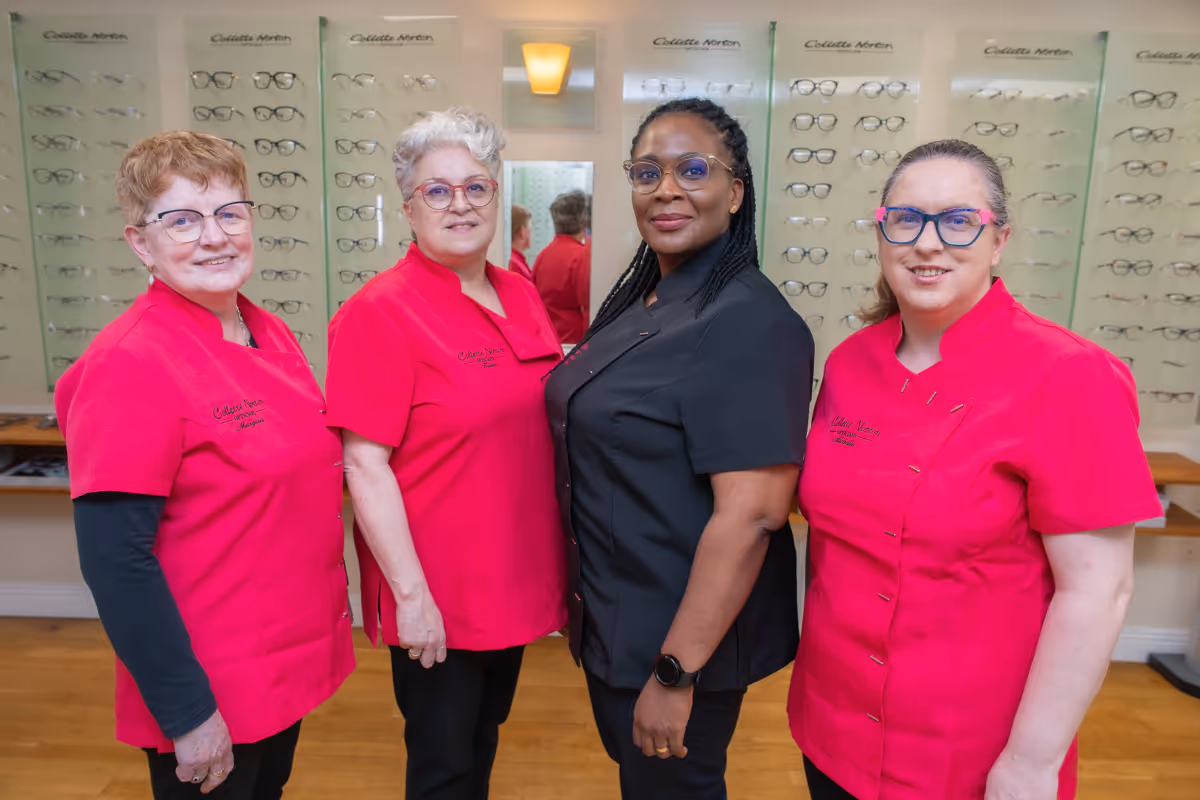 Four optometry staff members wearing pink uniforms pose together in front of eyeglass display cases.