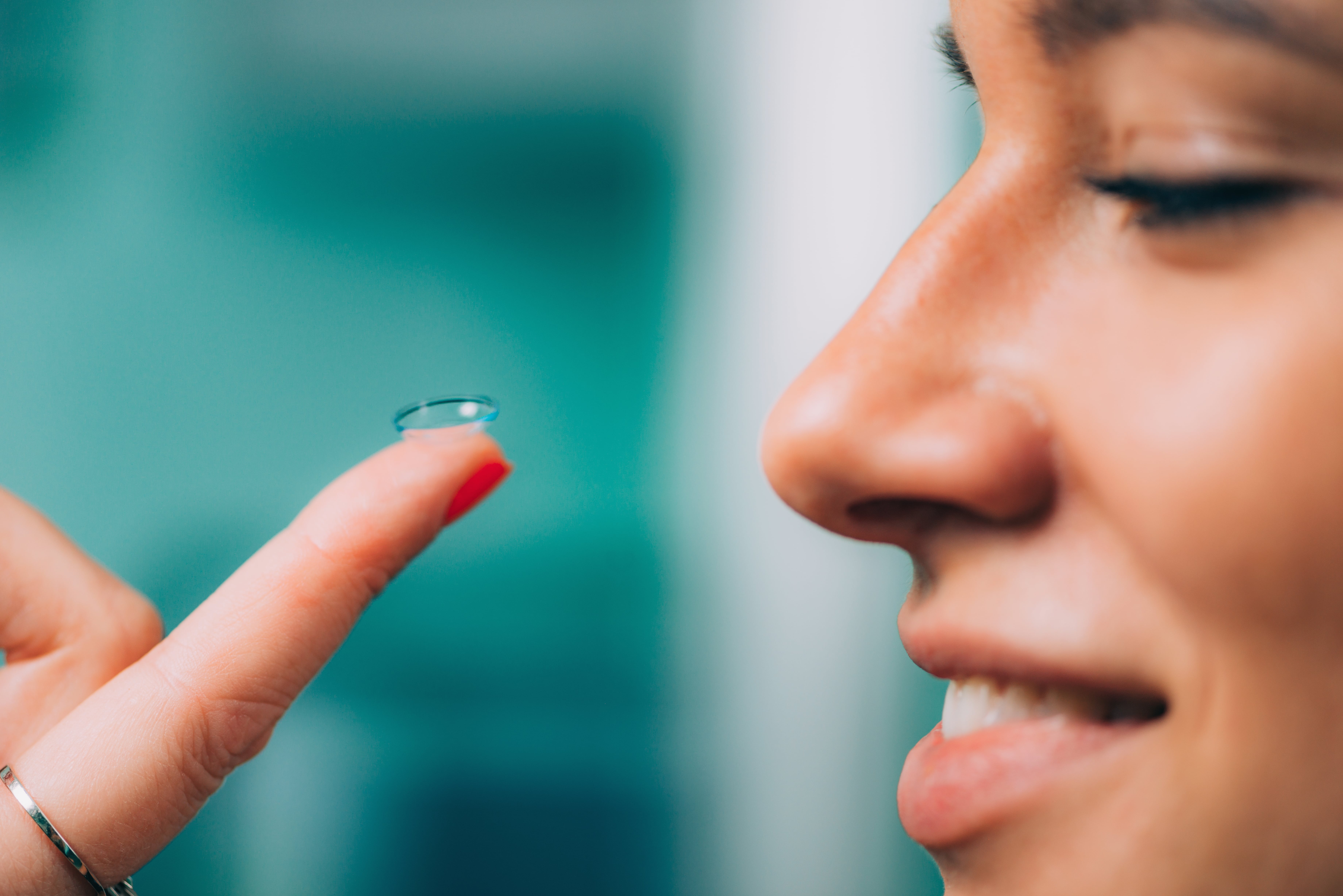 Person examining contact lens on fingertip against turquoise background