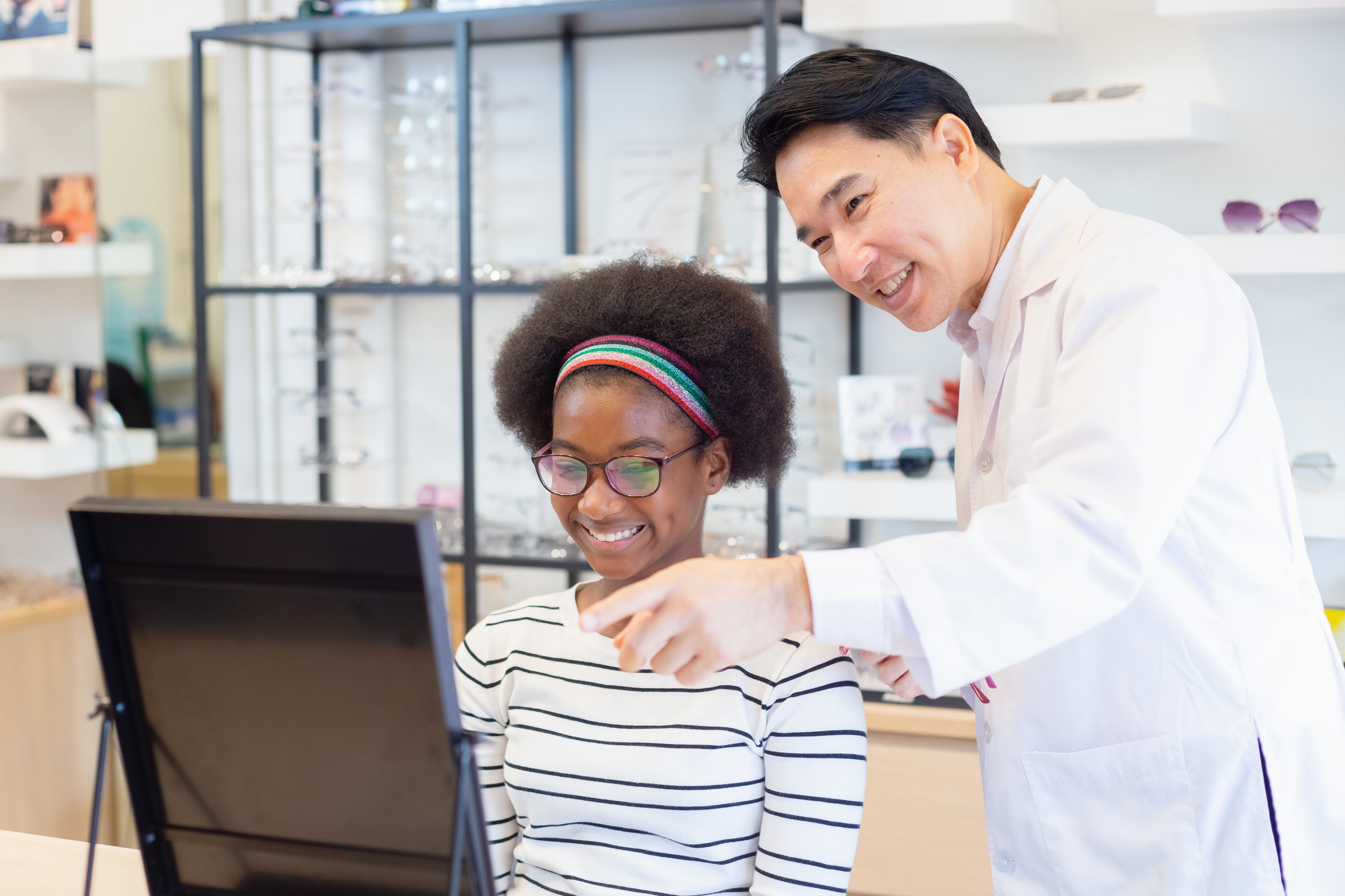 Female optometrist helping young girl try on eyeglasses in modern optical shop