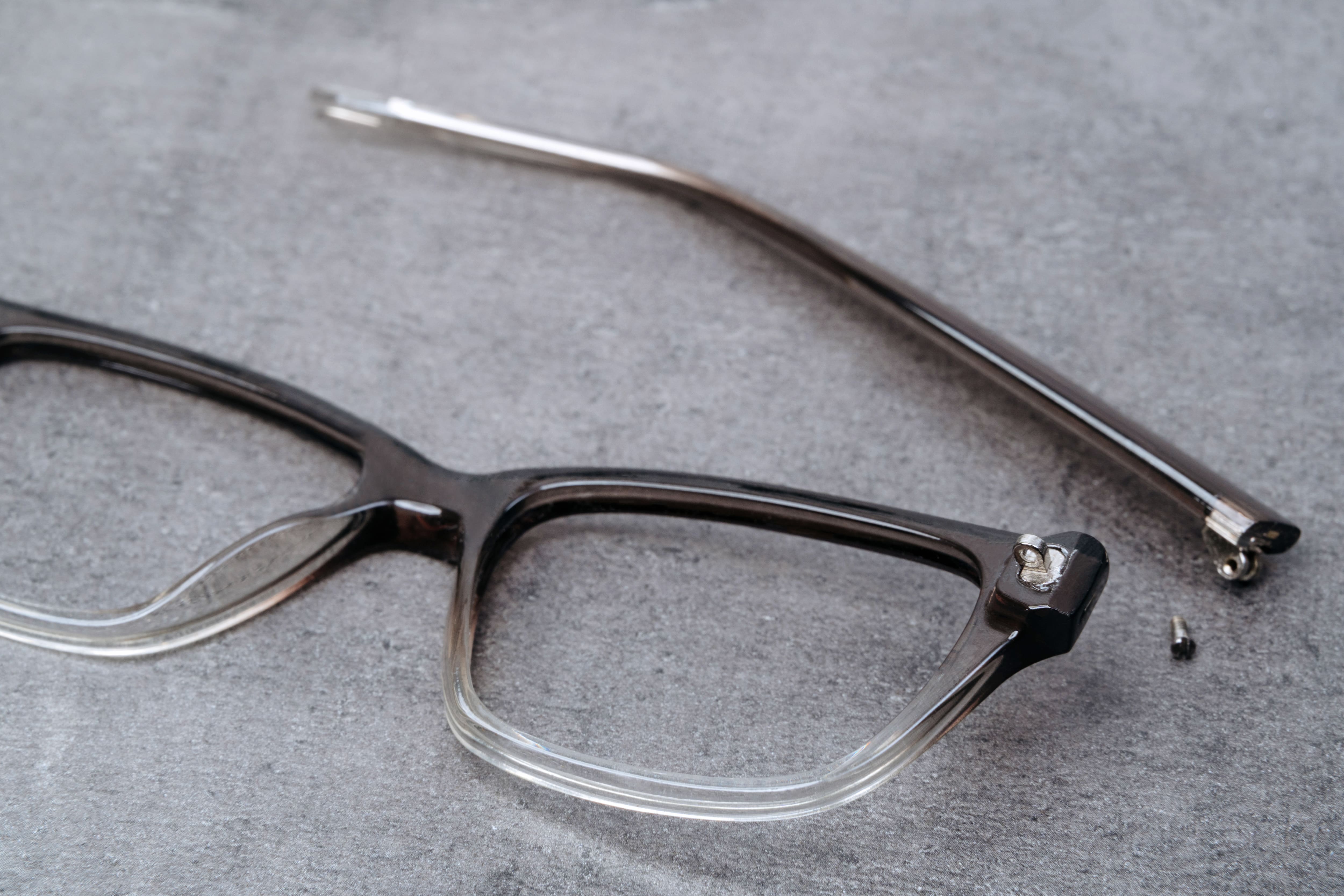Broken eyeglasses with cracked frame and detached screw on gray surface.