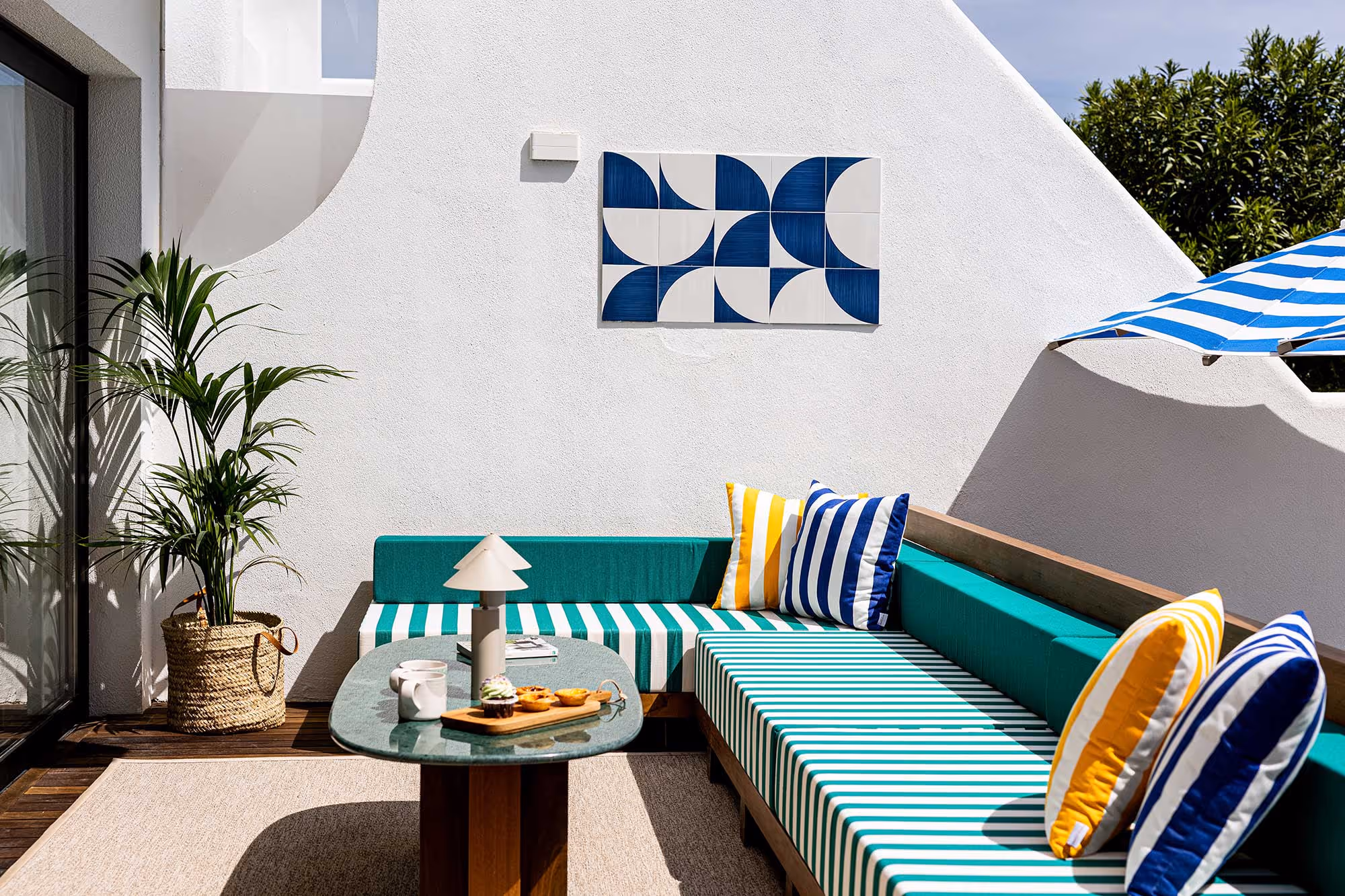 Sunlit outdoor seating area with green and white striped cushions, yellow and blue striped pillows, a green marble table with desserts, potted plant, and blue and white striped umbrella.