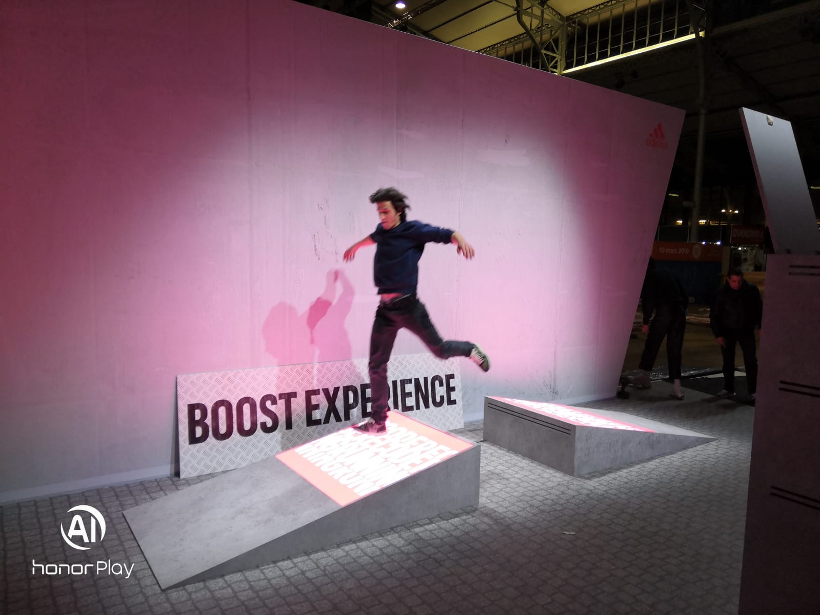 Person mid-jump on a concrete ramp in front of a pink wall with a 'BOOST EXPERIENCE' sign and Adidas logo.