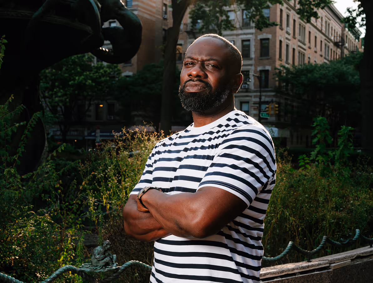 Man in striped shirt standing confidently in an urban park, with tall buildings and greenery in the background.