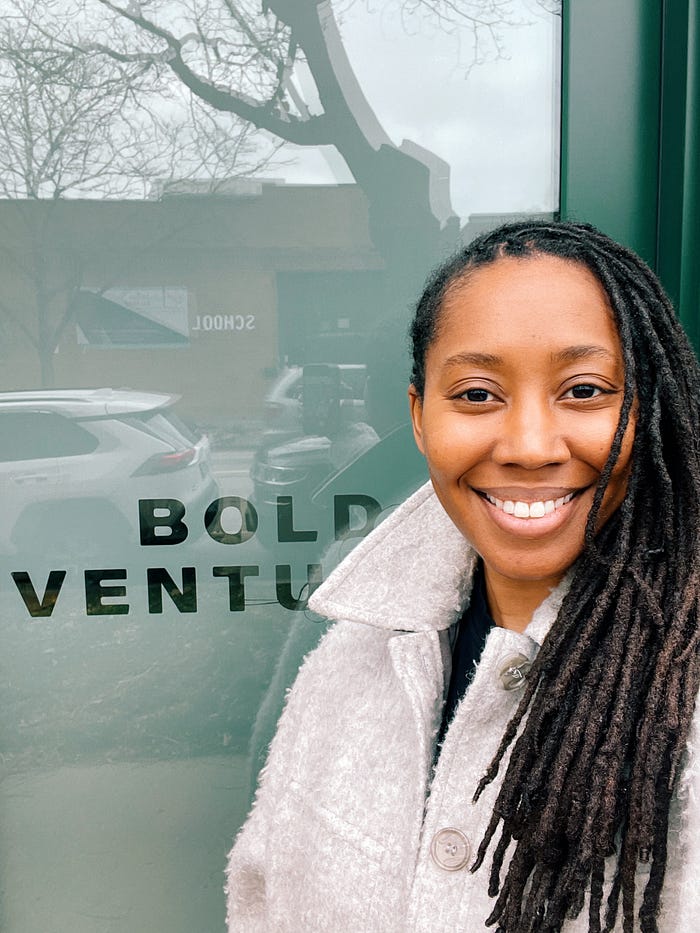 Jessyca Dudley stands in front of the Bold Ventures office, smiling. She is wearing a light-colored textured coat with a high collar. The glass door behind her reflects trees, parked cars, and a building with the word ‘SCHOOL’ visible. The words ‘BOLD VENTURES’ are printed in bold capital letters on the glass.