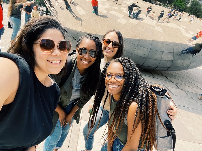 Melissa Gomez, Jessyca Dudley, Alysha Catalano, and Crystal Shaw of the Bold Ventures team pose for a selfie in front of The Bean (Cloud Gate) in Chicago. They are all smiling and wearing sunglasses. Melissa, on the left, is wearing a black sleeveless top. Jessyca, second from the left, is wearing a green jacket over a gray top. Alysha, third from the left, is wearing a beige top. Crystal, on the right, is wearing a green tank top and has long braided hair. The reflective surface of The Bean cap