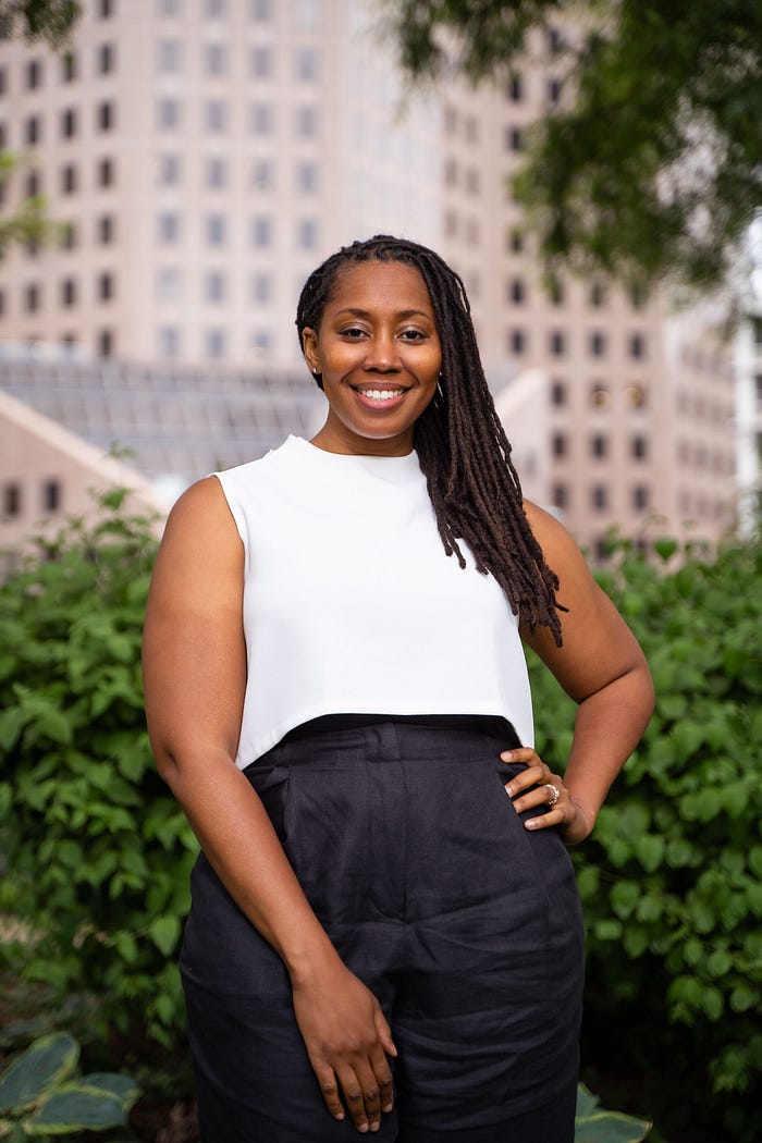 Headshot of Jessyca Dudley, a woman with long locs. She is wearing a white sleeveless top and high-waisted black pants. Jessyca is smiling confidently, standing outdoors with greenery in the foreground and a blurred building in the background.