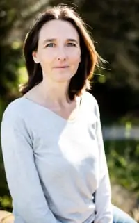 Frau mit schulterlangen braunen Haaren trägt einen hellgrauen Pullover und sitzt draußen im Sonnenlicht, leicht lächelnd in die Kamera schauend.