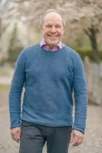 Älterer Mann mit grauen Haaren, blauem Pullover und Hemd darunter steht draußen, lächelt freundlich in die Kamera.