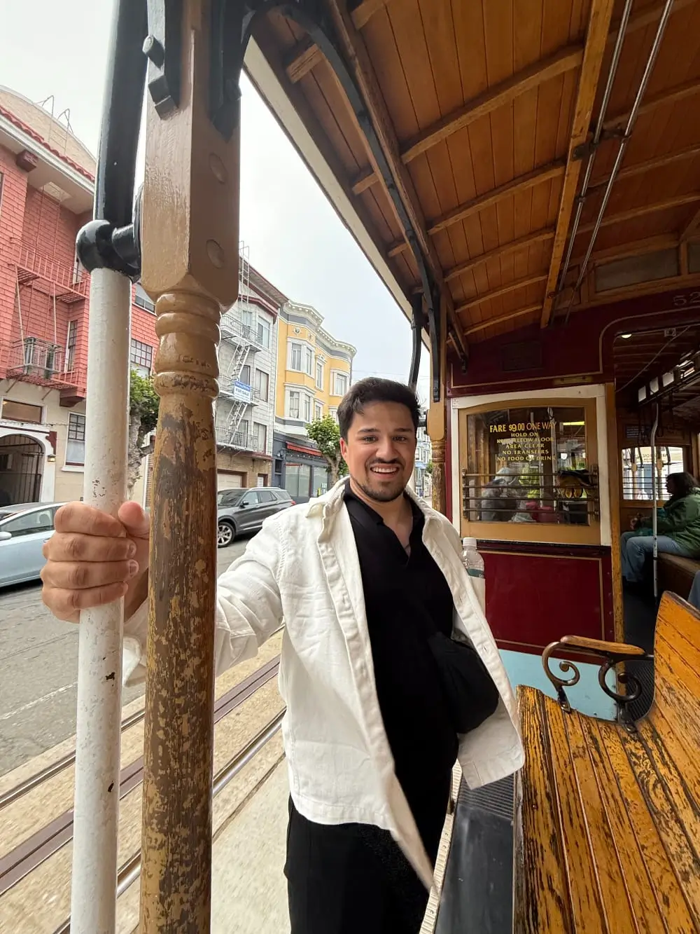 Mann mit weißer Jacke und schwarzem Hemd fährt in einer offenen Straßenbahn, lächelnd in einer Stadt mit historischen Gebäuden.