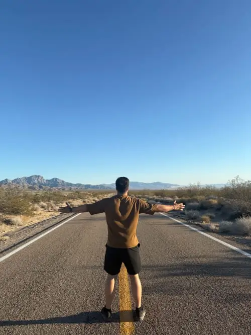 Mann steht mit ausgestreckten Armen mitten auf einer Landstraße in der Wüste, Berge am Horizont und blauer Himmel.
