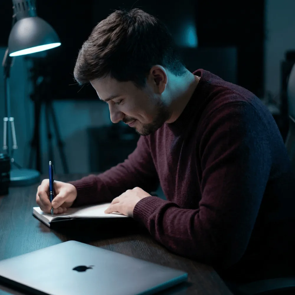 Ein Mann mit dunkelrotem Pullover schreibt nachts in einem Heft am Schreibtisch mit Schreibtischlampe und geschlossenem Laptop.