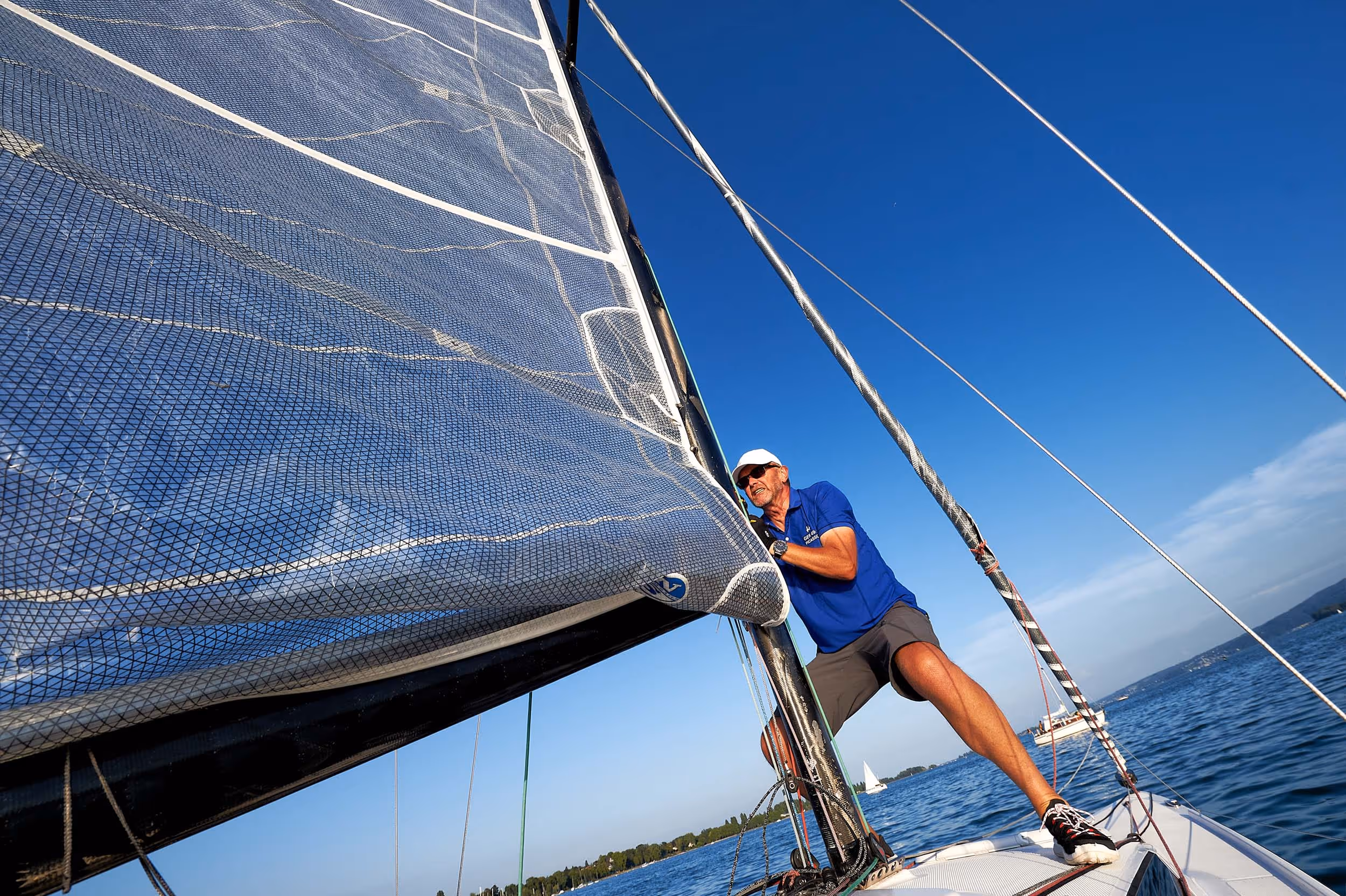 Martin beim Segeln. Er steht vorne am Mast und  zieht kraftvoll und dyna misch das Großsegel hoch