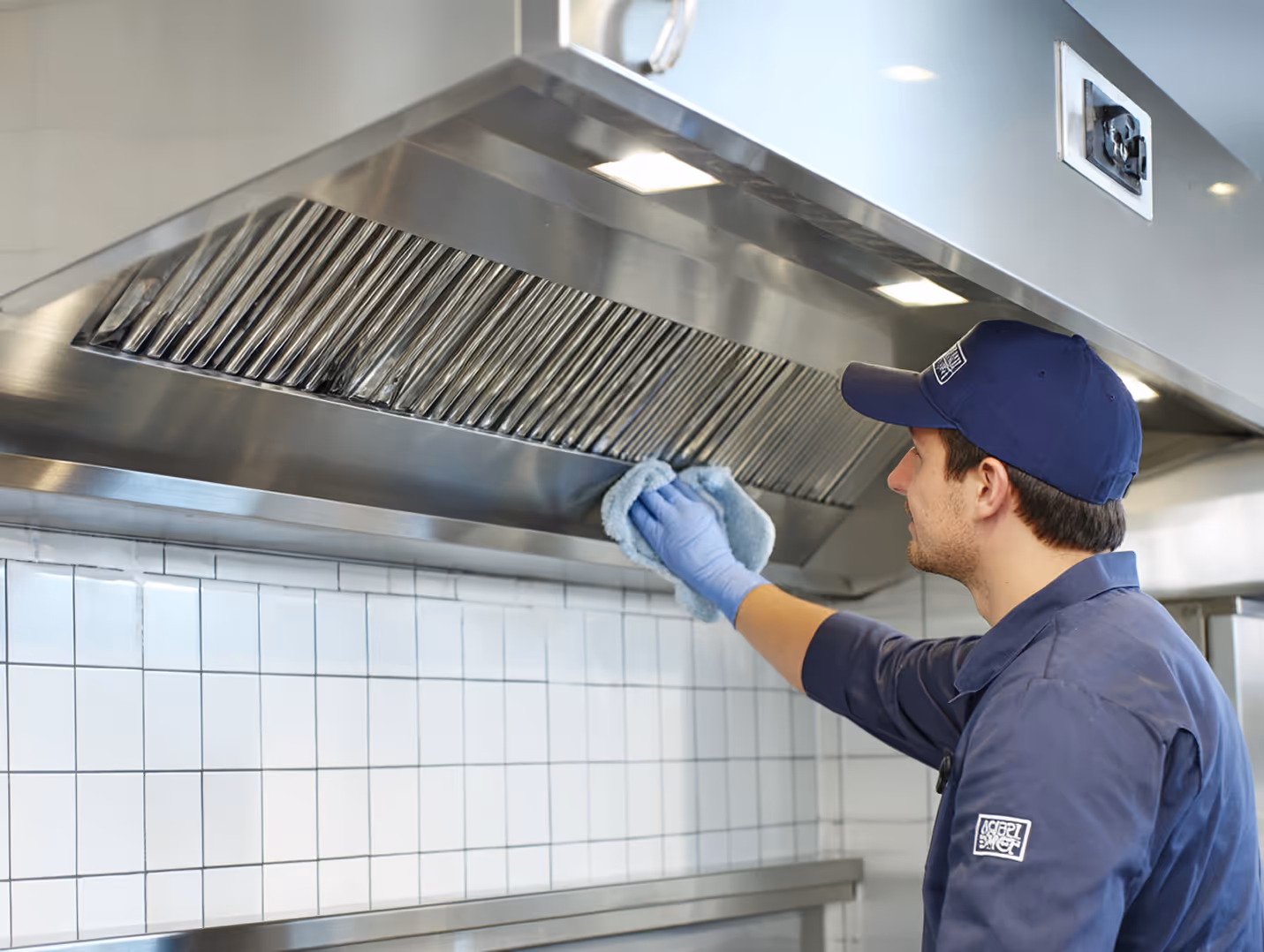 Reinigungskraft säubert professionell die Metallfilter einer Dunstabzugshaube in einer Küche mit Handschuhen und Tuch.