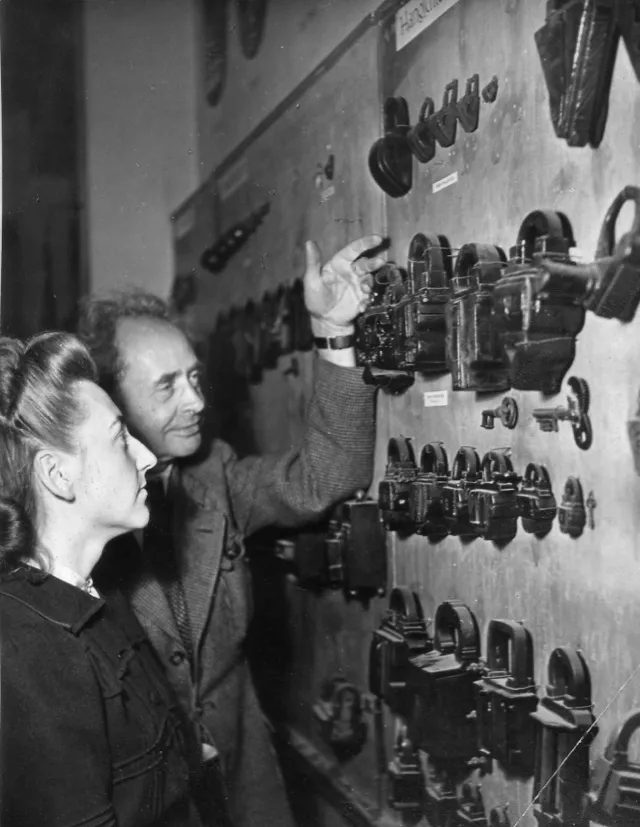 Auf einer schwarz-weiß Fotografie ist die alte Ausstellung des Museums zu sehen. Ein Mann zeigt auf ein Schloss, neben ihm steht eine Frau und schaut interessiert. 