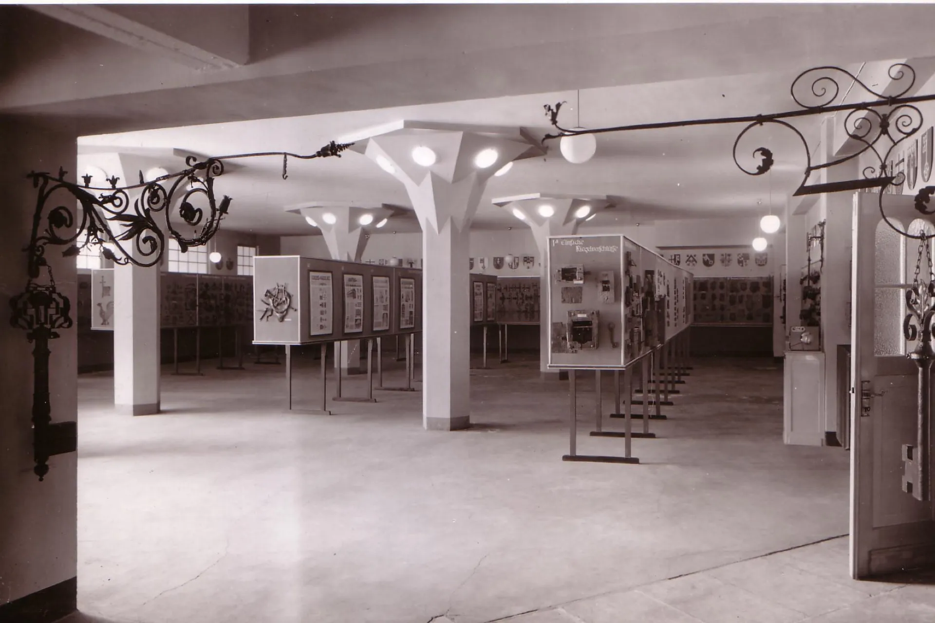 Auf einer schwarz-weiß Fotografie ist die alte Dauerausstellung des Deutschen Schloss- und Beschlägemuseums zu sehen. In einer Säulenhalle stehen mehrere Reihen mit Ausstellungsstücken. 