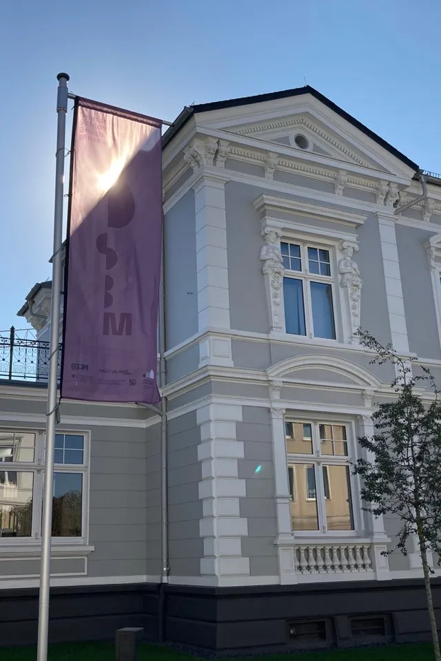 Eine Fotografie zeigt die Villa Herminghaus von außen, vor der eine Flagge mit dem Logo des Museums weht. 