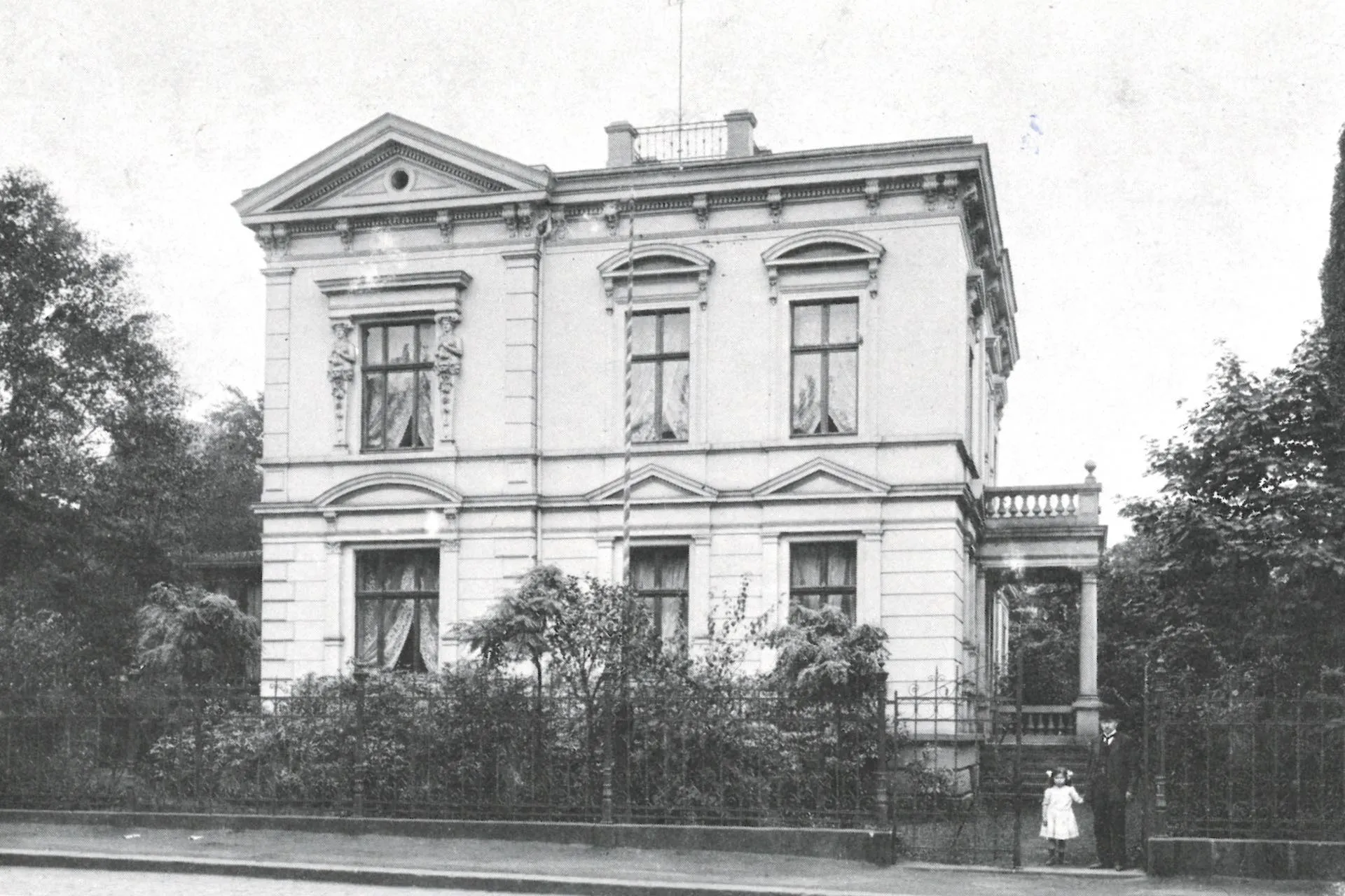 Eine historische Außenaufnahme zeigt die Villa Herminghaus in Schwarz-Weiß. Davor stehen ein Mann und ein Kind. 