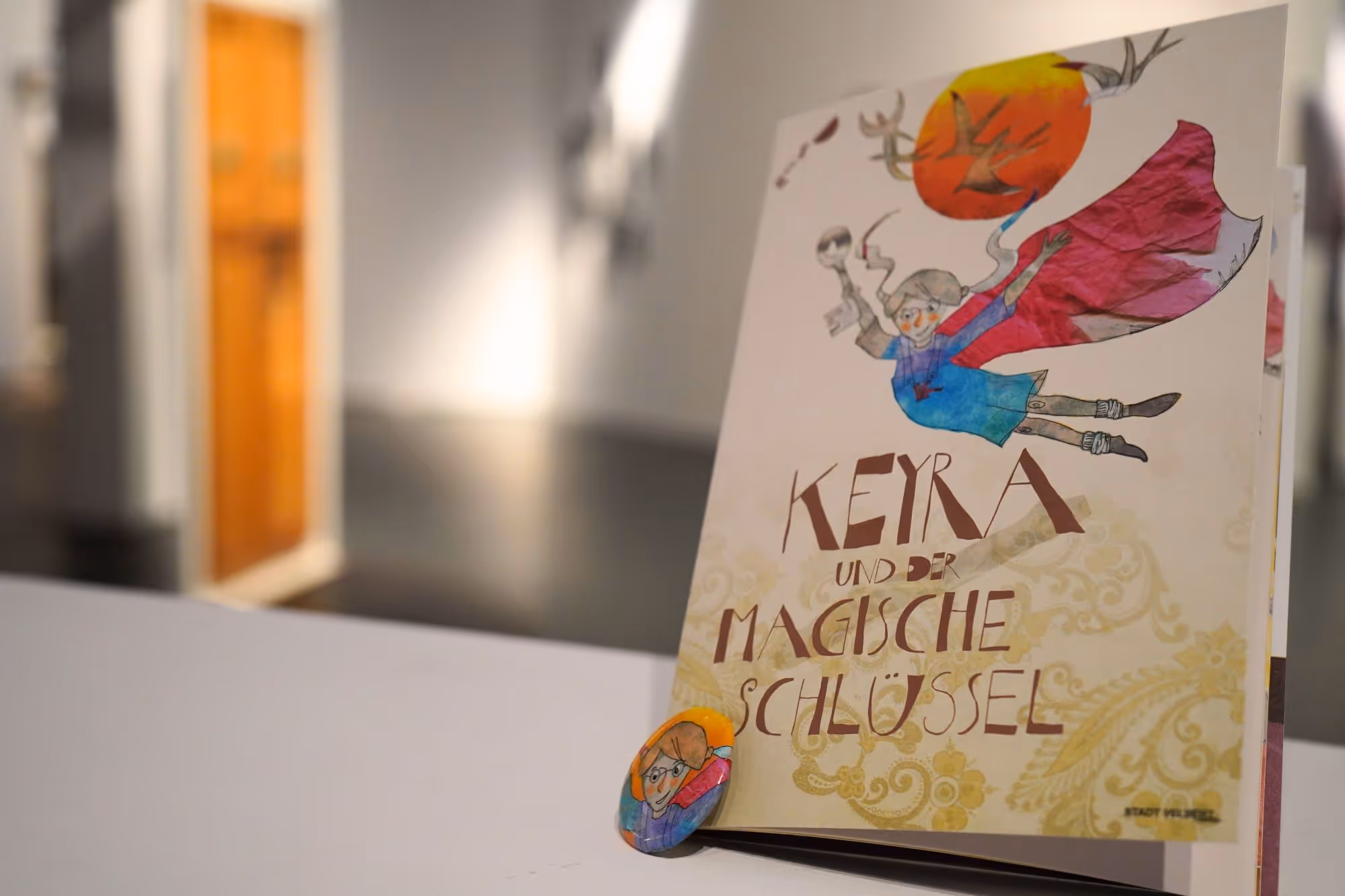 Ein Heft für den Kinderrundgang steht auf einer kleinen Mauer, dahinter ist die Ausstellung zu sehen.