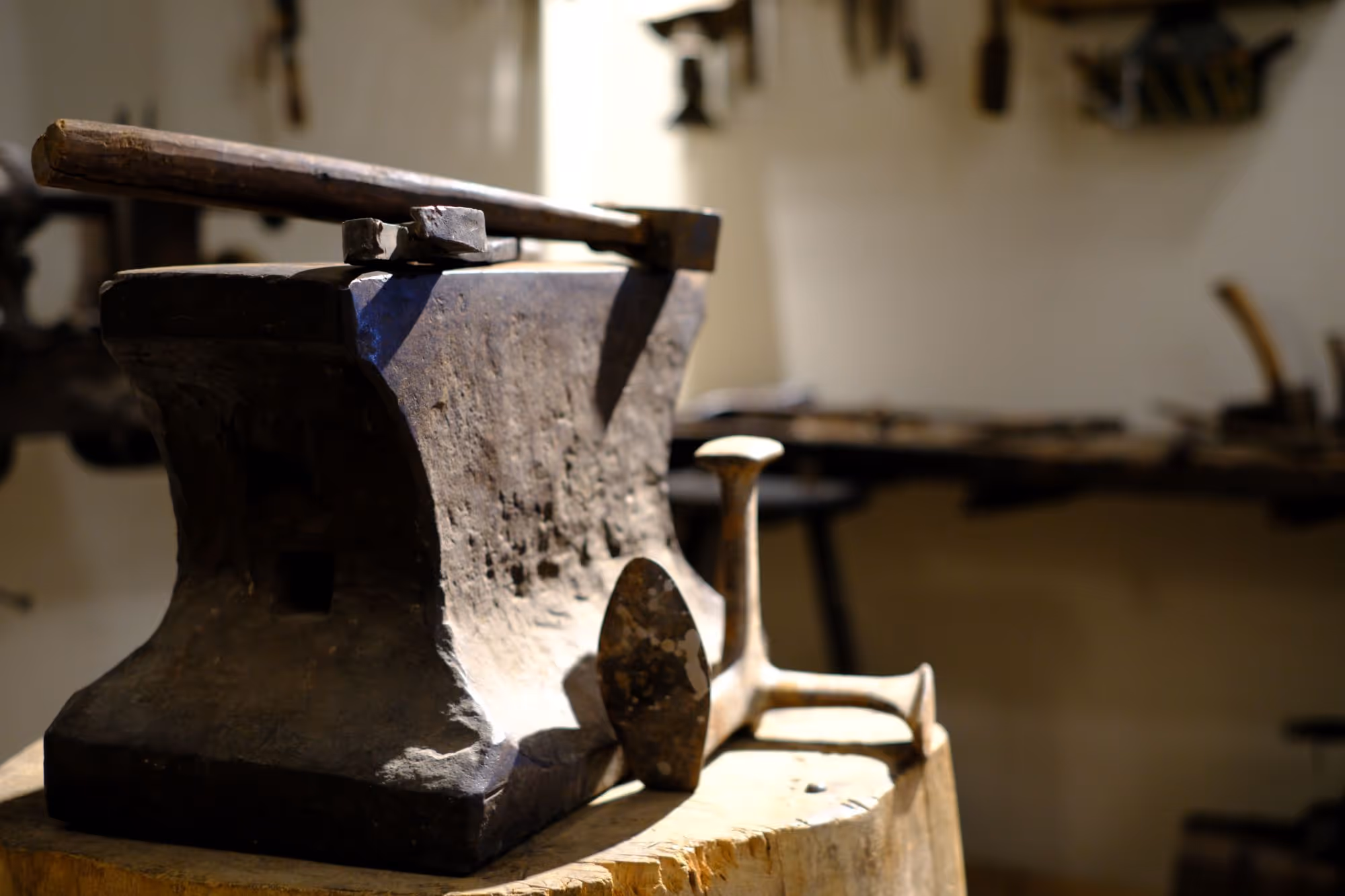 Ein Amboss mit einem Hammer steht im Vordergrund, im Hintergrund sieht man eine Werkbank mit weiterem Werkzeug.
