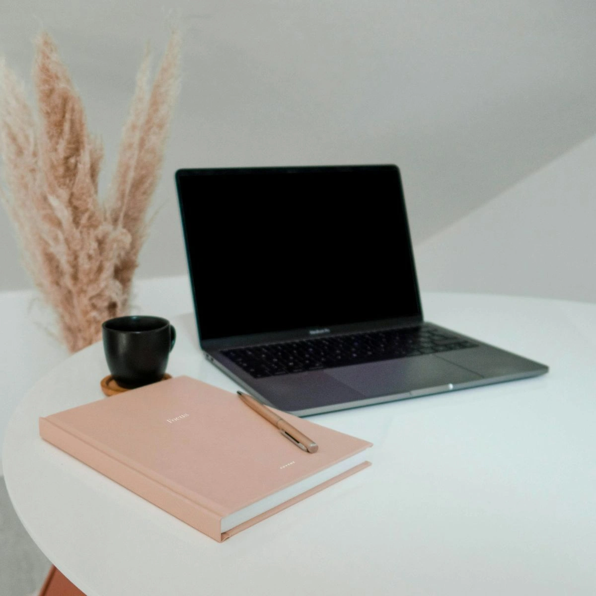 Laptop on white table beside a pink notebook, a pen, and a black cup of coffee