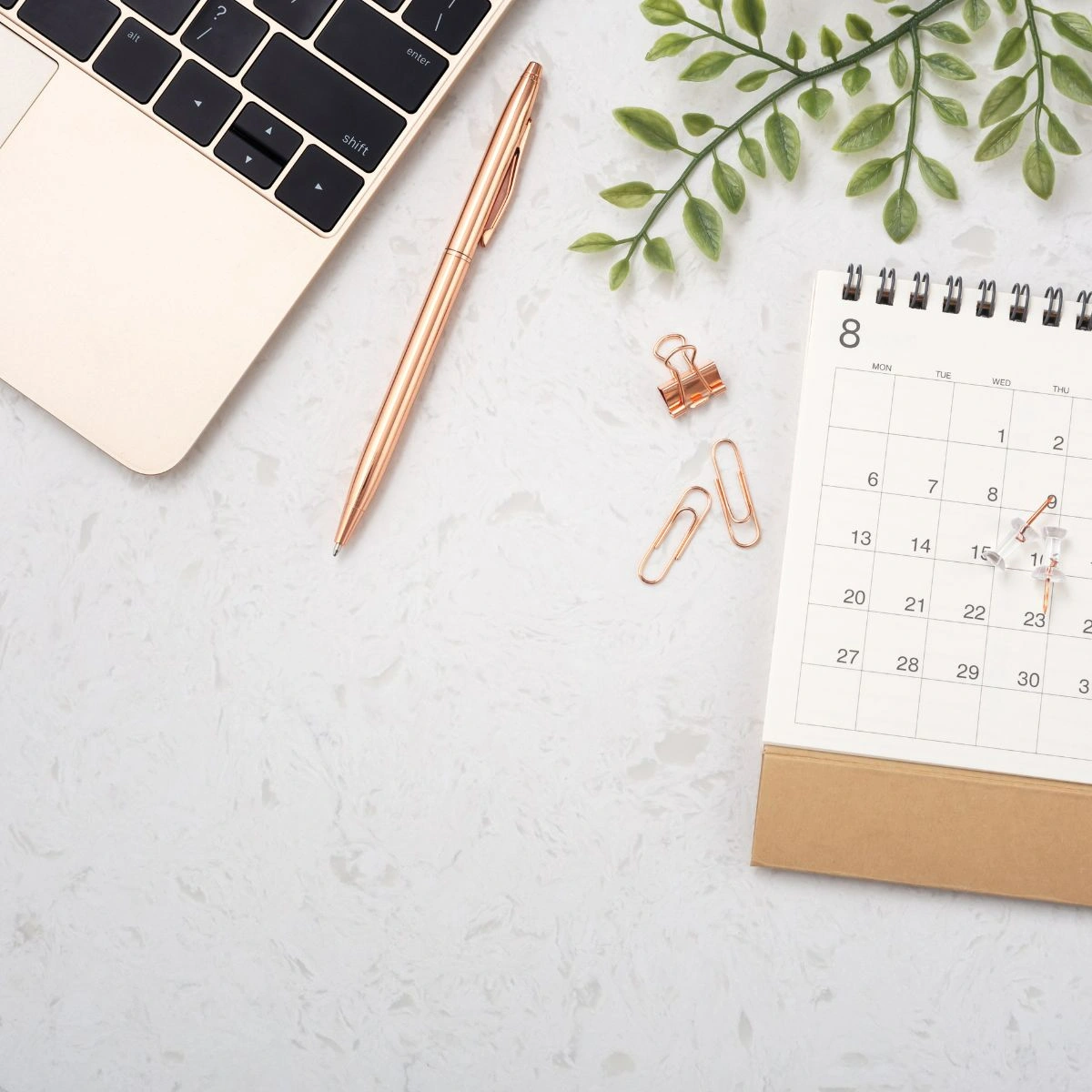 Flatlay of a laptop, a pen, calendar, a few clips and pins, and leaves