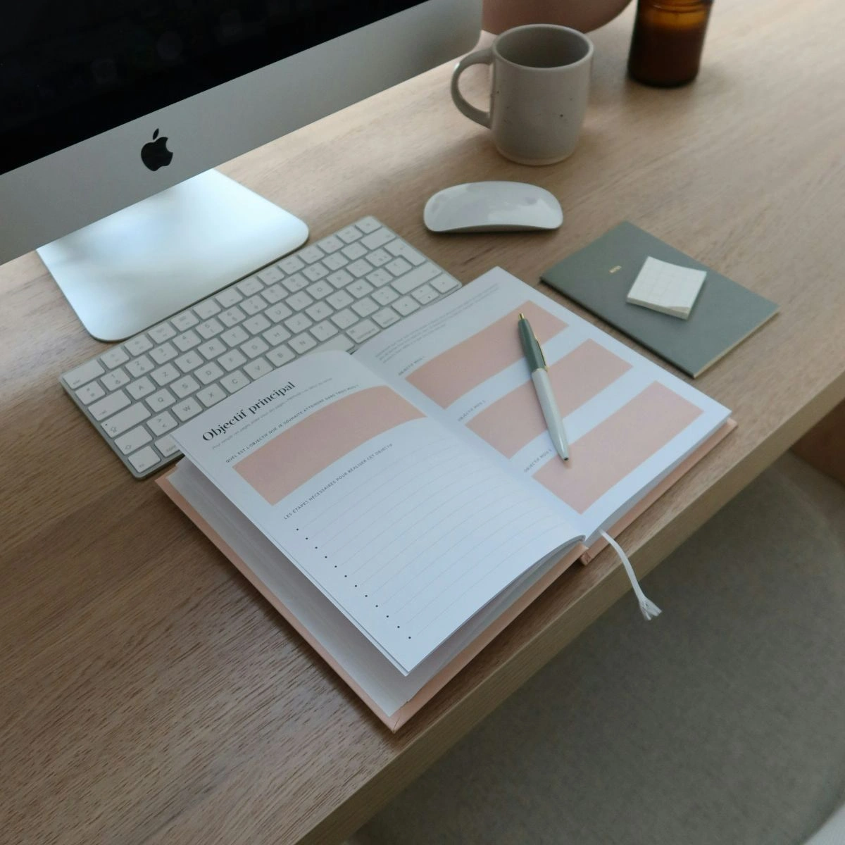 Close-up shot of a planner and a pen in front of an iMac and mug of coffee