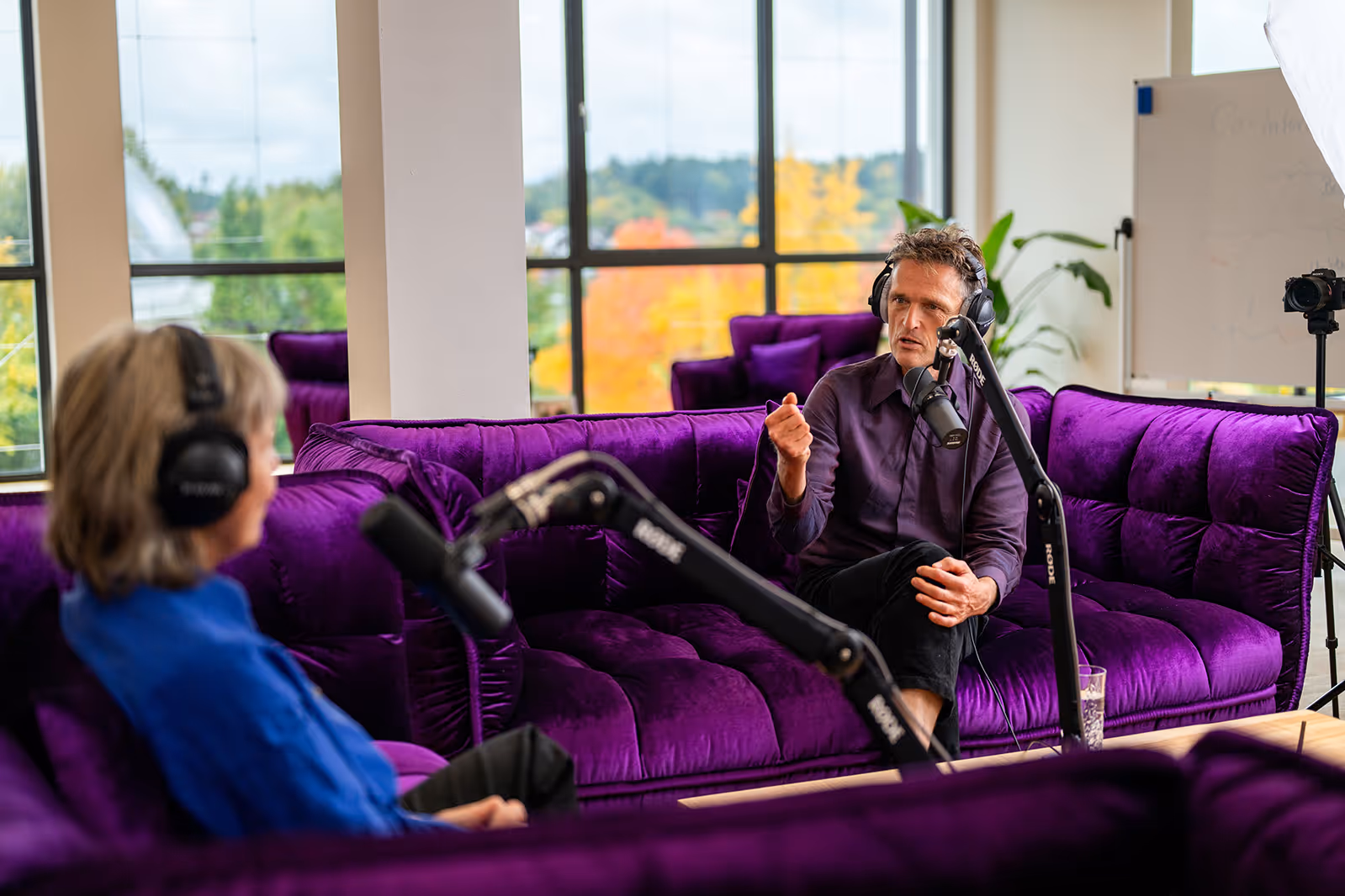 Gesprächssituation im Rahmen eines Deep-Democracy-Dialogs: Zwei Personen sitzen sich auf einem Sofa gegenüber, tragen Kopfhörer und sprechen in Mikrofone; die Atmosphäre ist ruhig, reflektierend und professionell.