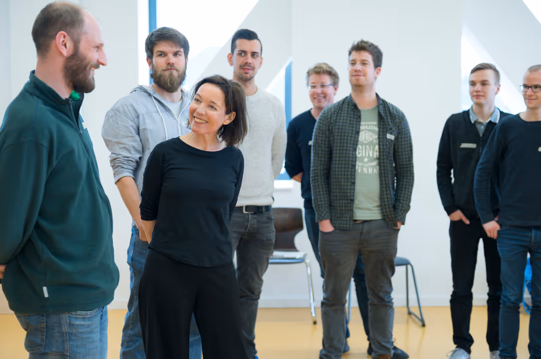 Christiane Leiste leitet einen Deep-Democracy-Workshop und steht im Austausch mit einer Gruppe von Teilnehmenden, die aufmerksam zuhören und in einem hellen Seminarraum im Halbkreis stehen.