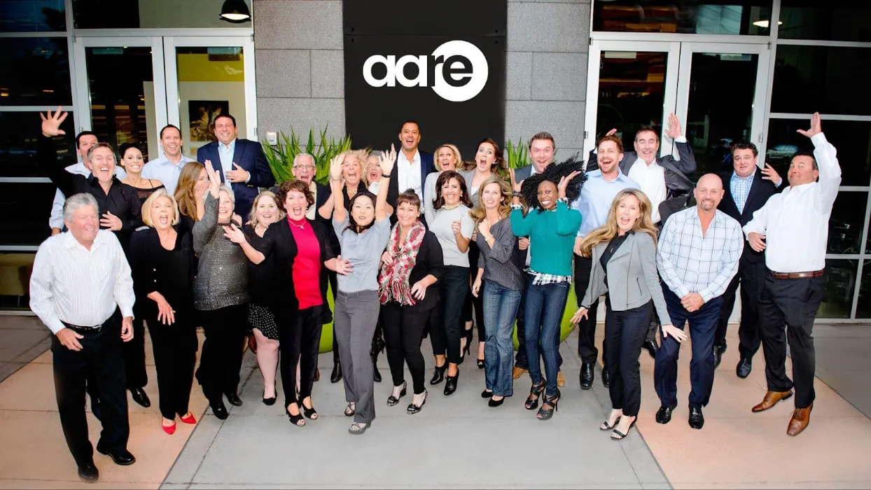 A worker on a ladder beside a modular home with large windows and a pitched roof being installed by a crane.
