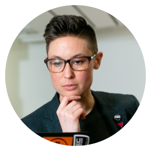 An image of a young working professional within a circular frame. They are wearing a trendy professional outfit and glasses whilst looking at a laptop screen in a meeting. They have a relaxed and confident expression.