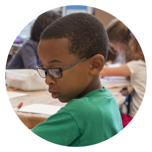 An image of a school pupil within a circular frame. They are in a class surrounded by friends, working in an exercise book.
