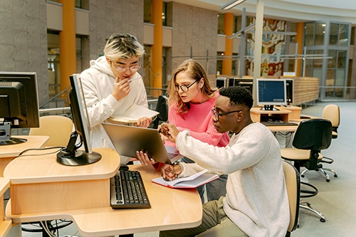An image of three university students gathered around a laptop to discuss their essential skills.