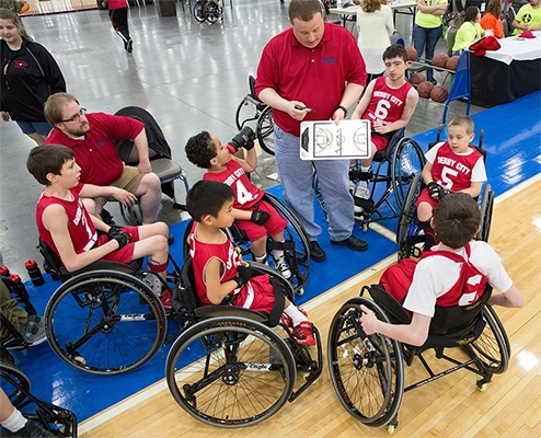 An image of  group of young wheelchair basketball players being given instructions by their coach in a team huddle.
