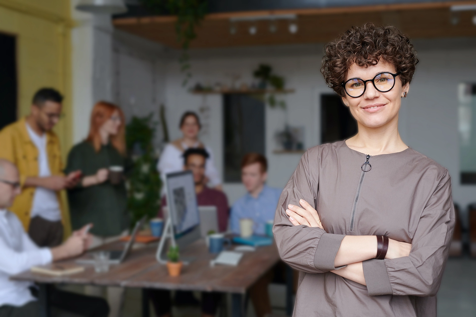 An image of a confident professional woman stood to the right of this image. She has curly hair and glasses and is wearing a modern chic outfit. She is stood tall and proud with her arms crossed and looking successful. Behind her is a blurred and darkened background of her colleagues working together on a project using a desktop computer.