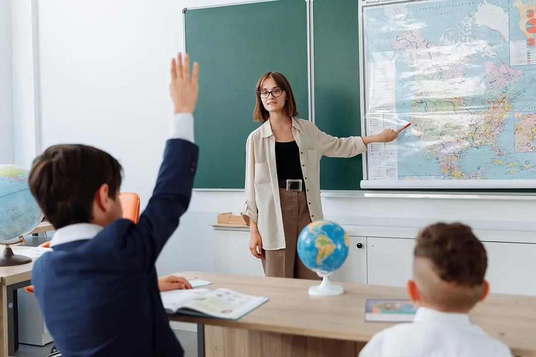 A teacher presents to her class. In the foreground a student raises their hand - they are wearing a blue school blazer with a white shirt collar. 