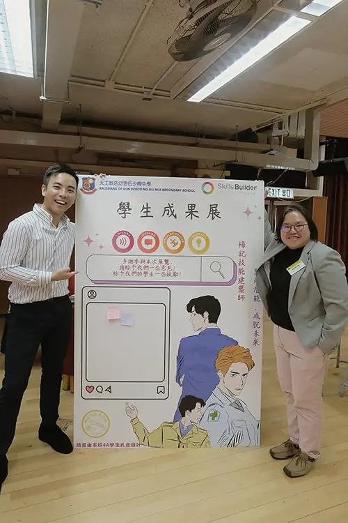 Marco Shek from Ednovators poses with a colleague in front of a poster board with the listening, speaking, problem solving and creativity icons on it. 