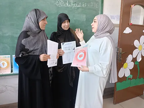 Three teachers in Lebanon stand and discuss with each other a lesson plan to incorporate speaking skills. They are wearing long dresses with grey and cream coloured hijabs. 