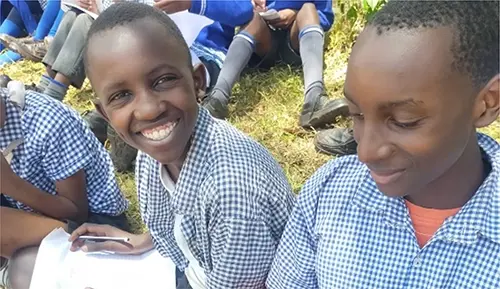 Two Kenyan boys in blue chequered school uniforms smile happily as they sit and study outside.