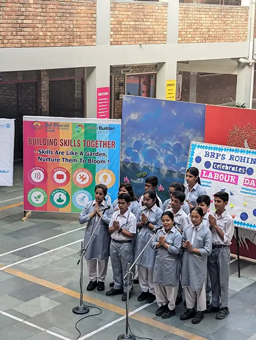 A group 12 Indian school students stand in a tight group as they deliver a presentation. Behind them is a large banner with the eight essential skills icons. 