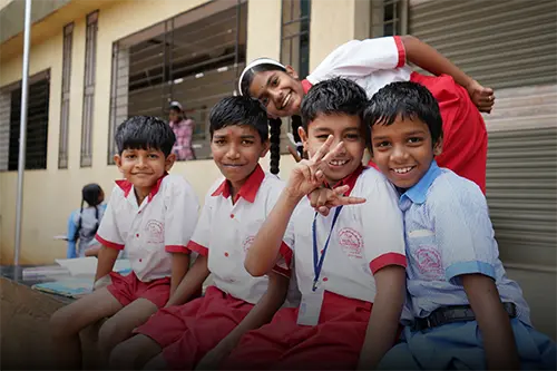 A group of young Indian schoolboys wearing school uniforms of white shirts and red shorts smile as they sit relaxing outside during their school break time.