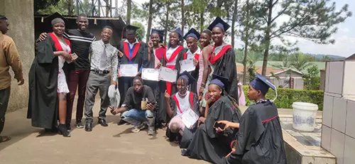A group of Ugandan college students wearing gowns and mortarboard hats stand and celebrate with their certificates on graduation day.