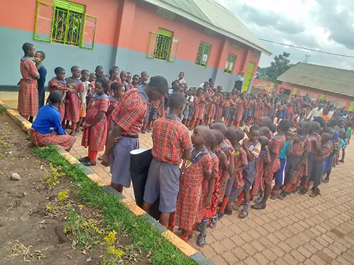 A group of young Ugandan school students stand in lines ready to return to lesson time, they are all wearing red tartan uniforms and are being instructed by older teenage students acting as the class monitors. 