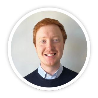 An image of John Sampson in a circular frame. He is waring a navy blue sweater with a light blue shirt collar, he has red hair and a warm, friendly smile.