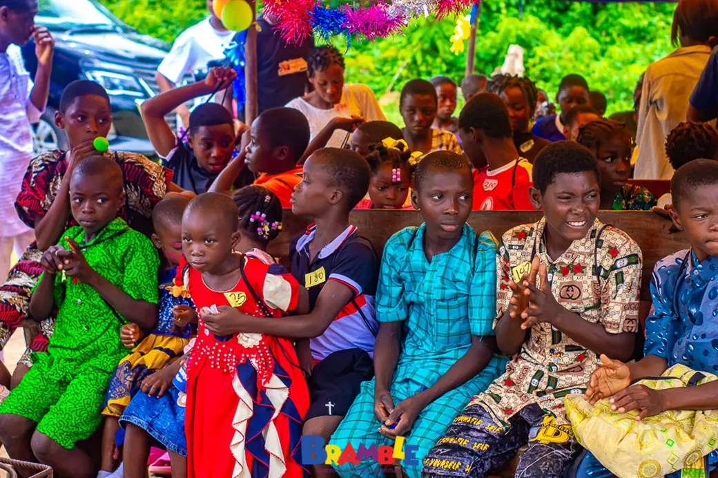 A group of 15-20 children aged around 7-10 sat for a lesson outdoors. They are wearing a mixture of brightly coloured shirts and traditional African dress.
