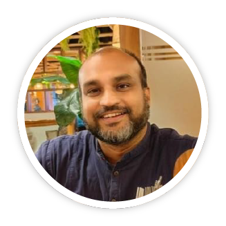An image of Pavan Kumar Kolli, an Indian man with a short beard, a joyful, kind smile and is wearing a dark navy blue grandad shirt