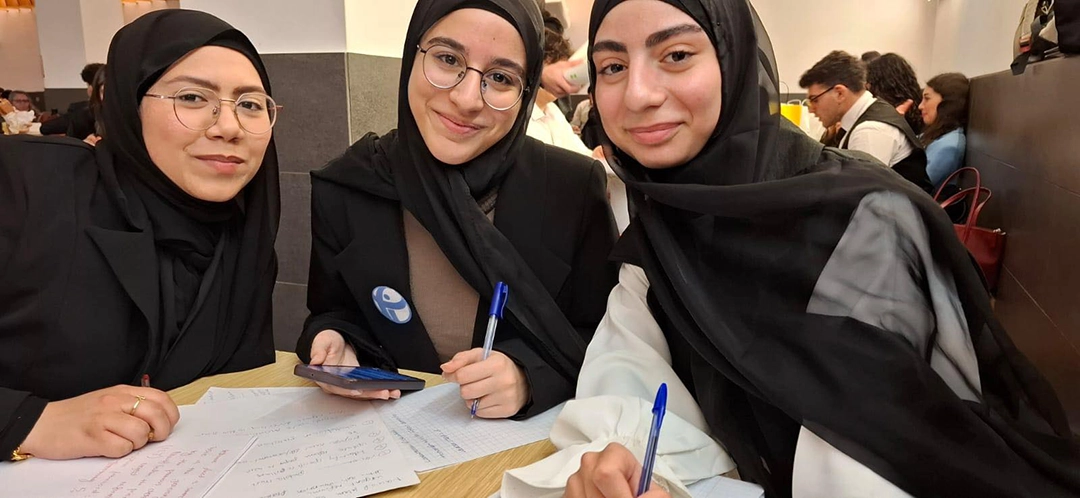 Three Lebanese girls in their senior year smiling for the camera as they revise for their final exams. They are smiling warmly and excitedly. They are wearing their school uniform of black hijabs and blazers.