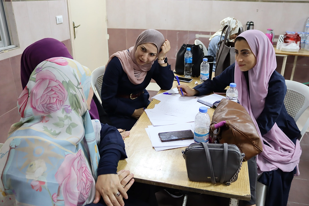 Three female school teachers from Imam Al-Hassan school sat together around a table discussing lesson plans for the upcoming term. They are smiling and chatting, engaged deeply in conversation. They are wearing smart-professional outfits with a mixture of colourful hijabs in peach, pink, purple and floral patterns.